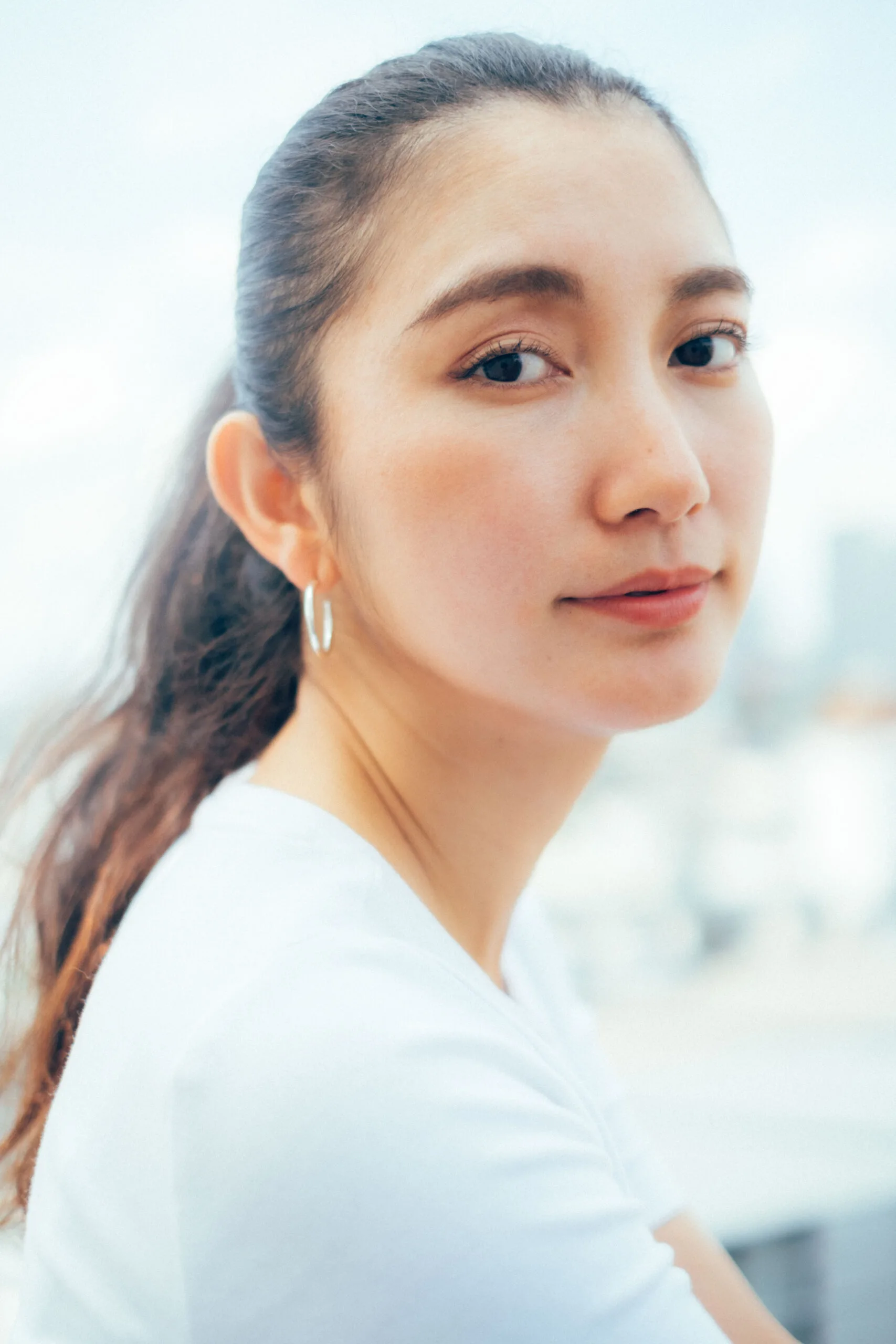 A person with long dark hair tied back, wearing a white top, looking at the camera. They are outdoors against a blurred cityscape background.