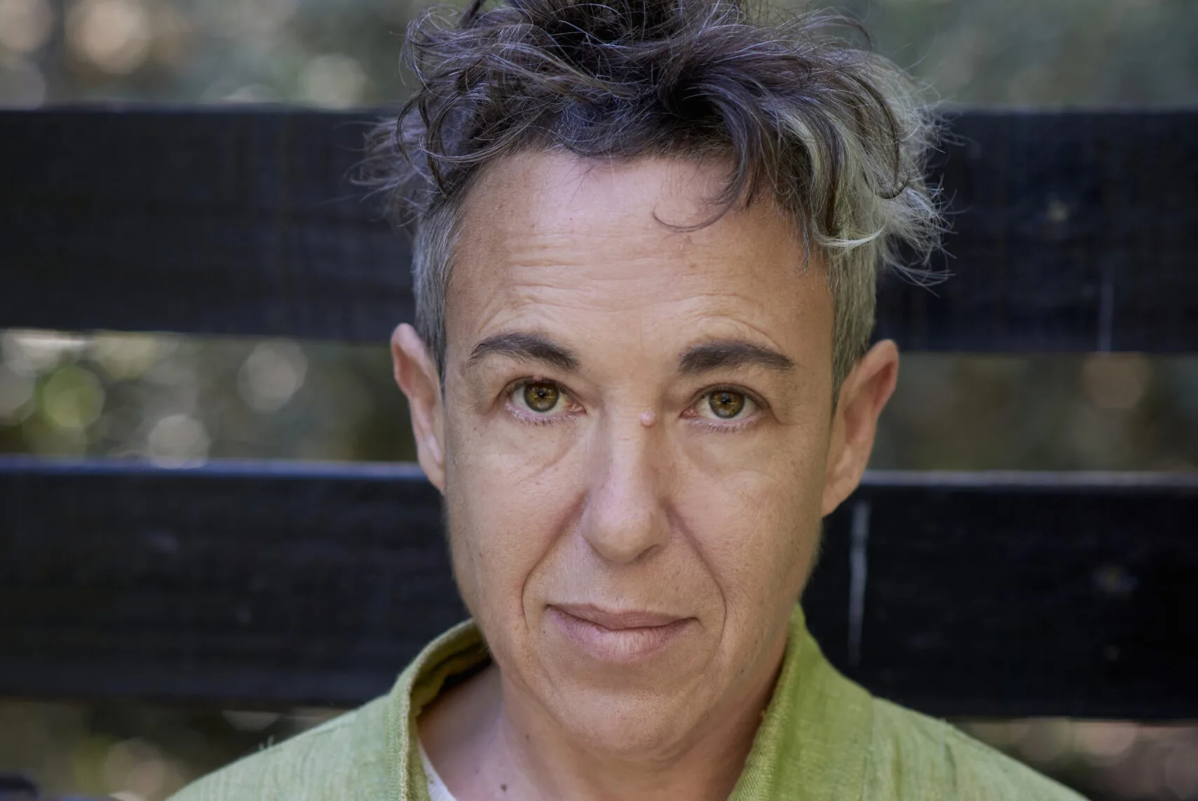 A person with short, tousled hair, graying at the roots, stands against a dark wooden background. They wear a light green jacket over an orange shirt with a striped neckline, looking into the camera with a neutral expression.