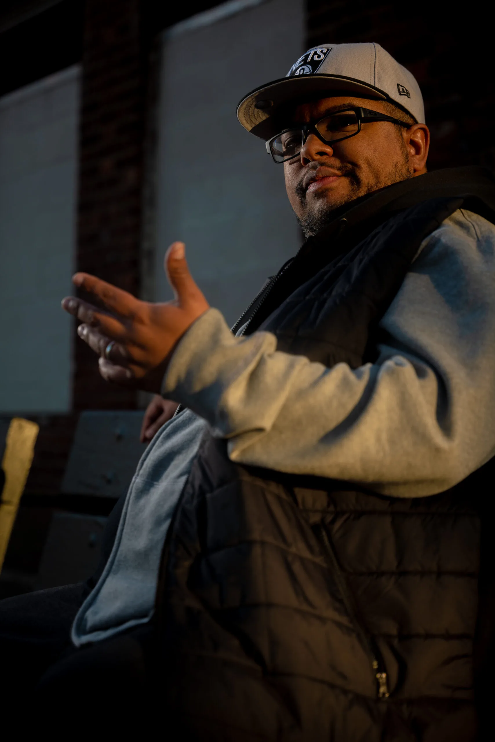A man wearing glasses, a cap, and a black vest over a gray hoodie sits on a bench, gesturing with one hand. The lighting casts dramatic shadows on his face and the background.